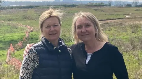 Laura Coffey/BBC Two women, one with a blue gilet and white top with black spots, and the other with a black top. Both have blond hair - Ms Whitcroft's (left) is tied back while Ms Croft's is untied. There is a field behind them with red fencing to the left.