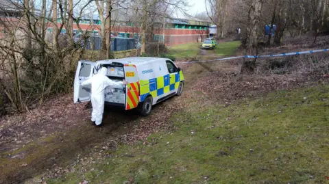 A man dressed in a white forensics outfit opens the two back doors of a police forensics van. It is parked in the middle of a wooded area, with blue and white police tape attached to some of the trees. Another police car and a building are visible in the background.