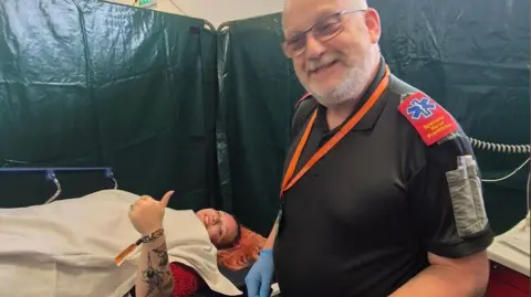 Keith has white hair and is wearing a black top with red medical shoulder pads. He is wearing blue medical gloves and is standing next to a young woman who is lying down on a medical bed under a white blanket. She has pink hair and is holding up her thumb