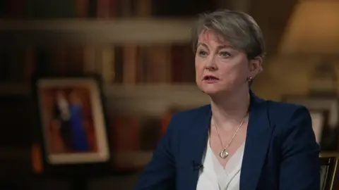 Home Secretary Yvette Cooper looks to the left of the camera. She wears a navy blazer, white top and silver necklace. 