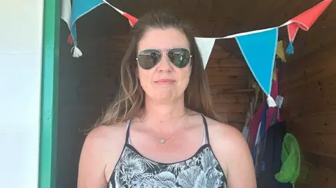 A head and shoulders shot of Sally Thomas, who is standing in the doorframe of her beach hut. She had long brown hair with lightened ends, is wearing wire-rimmed sunglasses and is smiling slightly. She wears a spaghetti-strap black and white top with a pattern of fronds on it, and what looks like straps from a swimming costume underneath. She also has a thin silver chain with four or five rings looped on at the front of it. Immediately behind her head is turquoise, white and red bunting. Items for the beach can be seen on one side of the cabin in the shade.
