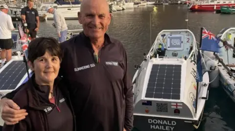 Nicky & Bob Parr The couple are standing on a dock in purple tops next to their boat. Mr Parr has his arm around his wife.