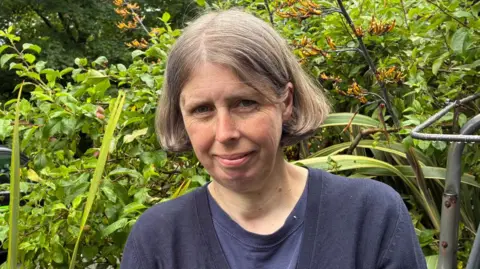 Sharon Morgan, a woman aged 50 standing in the front garden of her house. She has honey blonde bobbed hair and is wearing a navy top and navy cardigan.