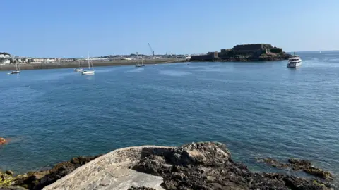 Havelet Bay - A picture of the blue sea, with a few sail small boats with their anchor dropped to the left aqnd a large grey yacht to the right. In the background Castle Cornet is pictured, a granite fortress, with the Castle Emplacement rolling down the left. 
