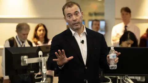 Will Lewis stands in a suit and white shirt, gesturing as he speaks to people in a newsroom.
