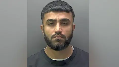 Hertfordshire Police A mugshot of Galmne, wearing a white t-shirt under a black sweater. He has a black beard and moustache and short dark hair.