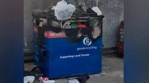 Tendring District Council A blue recycling bin with rubbish pouring out of it. It is also burnt at the top so the blue bin has become discoloured and turned black.