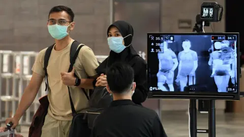 ROSLAN RAHMAN A couple, wearing protective facemasks amid fears about the spread of the COVID-19 novel coronavirus, walk past a temperature screening check at Changi International Airport in Singapore on February 27, 2020