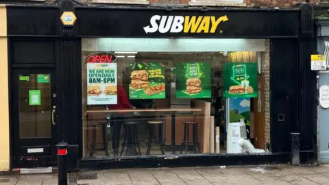 A black-painted building with a large Subway logo above a window stretching the length of the wall. There are posters of offers displayed in the window with a neon sign reading 'open'.