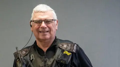 Staffordshire Police Police officer Kevin Lee is stood against a grey background. He is wearing glasses and black police uniform with a body-worn camera, police radio and other police equipment attached.