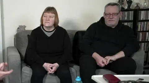 Denise and Ray sitting in their living room on a grey settee