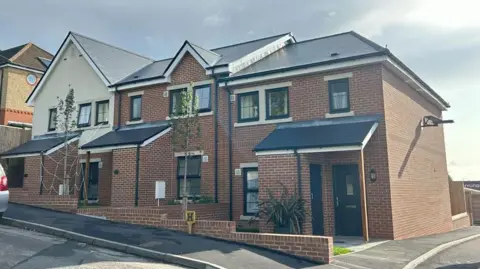 A row of three terrace houses. Two of them are brick while the third is half brick and half white rendering. They all have large porches and black windows on both floors.