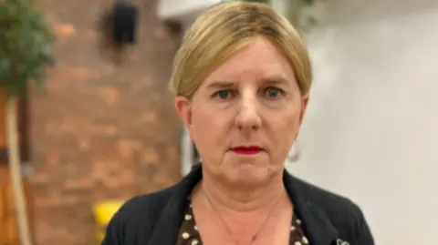 A headshot of Kaz Self standing in a back yard with a brick wall in the background. She is staring to camera and is wearing red lipstick, a black jacket and a brown and cream polka dot dress