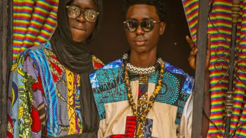 Two men in multi-patterned colourful patchwork clothes look at the camera framed by a doorway. One is wearing a black headscarf and glasses, the other is in sunglasses and has beaded necklaces around his neck - in Diourbel, Senegal, April 2024