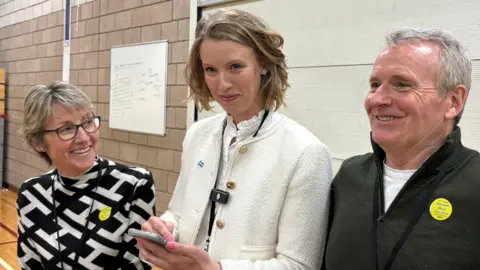 Hannah Goodlad, SNP winning candidate, smiling, in between party representatives.
