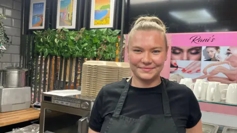 BBC Leah Williams, wearing a black t shirt and apron and standing in a cafe with a microwave, pots and cups behind her