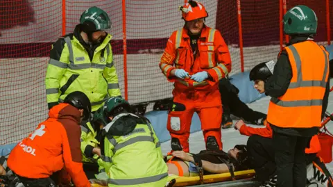 Hertfordshire County Council Rescue workers in helmets and high‑visibility jackets treat a trauma mannequin in a simulated environment. The mannequin is lying on a stretcher beside a fenced ski area, while others assist nearby.