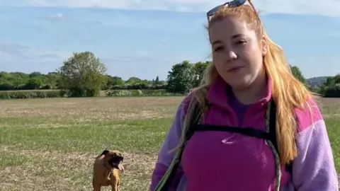 A woman with long strawberry blond hair has a pair of sunglasses on her head and is wearing a pink and purple fleece top. She is standing in a field with a dog in the background