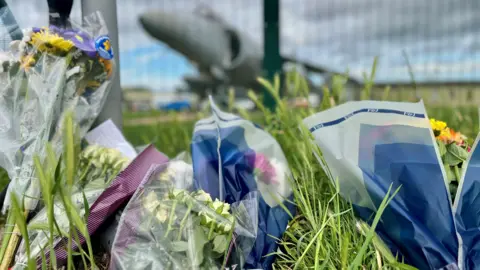 Bouquets of flowers left outside RNAS Yeovilton. The flowers are on a grass verge. A fighter jet can be seen in the background behind a green mesh fence, although it is blurred.