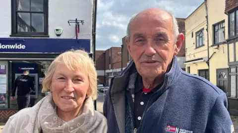 Holly Nichol/BBC A woman and man standing side by side in a street. The woman has short blonde hair and is wearing a cream patterned scarf over a cream fleece. The man has short white hair and is wearing a navy fleece jacket over a zipped up jumper and shirt. Behind them is the corner of a street with part of a navy sign saying Nationwide and a man is standing in front of a cashpoint. A street stretches off behind them. 