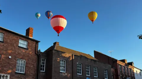 Four hot-air balloons flying quite low above a row of terraced houses. The sky is light blue and completely clear