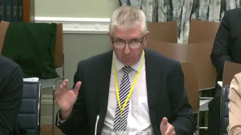 NI Assembly Man wearing a suit and tie, glasses and a yellow lanyard sitting at a desk in a Stormont committee room. There are some empty chairs behind him