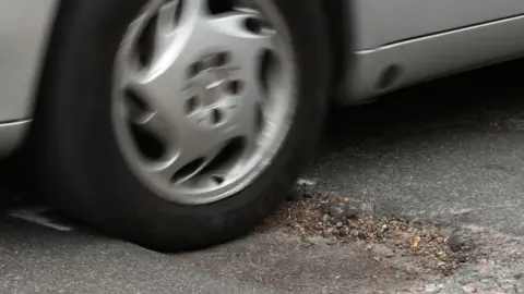 A close up of car wheel hitting a pothole in the road. The car is silver. 