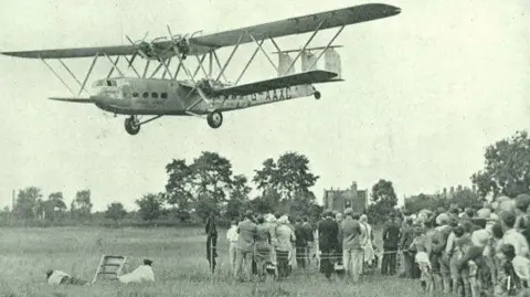 A black-and-white photograph of a Hercules aircraft approaching Brockworth airfield in 1932. The plane is still airborne, descending over a wide grassy field. On the right side of the image, a crowd of spectators stands with their backs to the camera. Two people dressed in white are lying on the grass in the foreground, apparently trying to get a better view. The scene captures the anticipation of the landing.