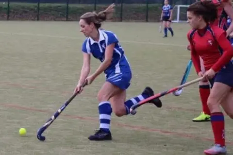 Clwb Hoci Rhuthun Claerwen running with her hockey stick stretched in front of her next to a yellow hockey ball. She is wearing a blue hockey kit. To the right is a player wearing a red hockey kit with her hockey stick in front of her.