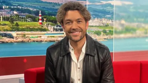 Emrhys Cooper sat on a red sofa. He is wearing a black leather jacket, white shirt and brown trousers. The sofa is red. There is a backdrop of Plymouth Hoe on the screens behind him.