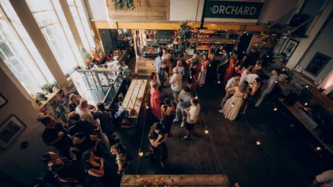 The Orchard Bristol The camera looks down on a busy cafe floor, crowded with people, with light shining in through big church windows. This is the Orchard Cafe, in St George in east Bristol. 