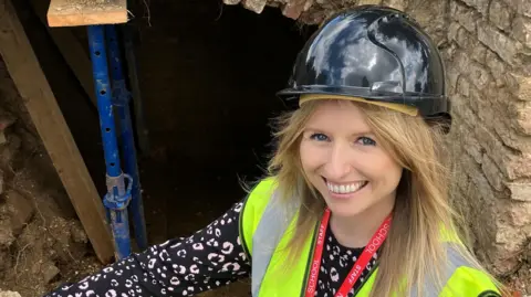 A woman in a hard hat and a high-vis yellow vest proudly squatting in front of an opening into a tunnel. She is smiling.