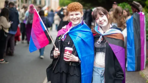 Marie-Claire Parker Photography Two young women stand among the crowd at the Pride event wearing colourful flags as capes. One woman has her hand around the other's shoulder.