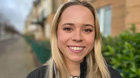 Phoebe is stood in a street, smiling at the camera, she has long blonde hair and is wearing a black coat.