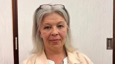 LDRS Sarah Jane Colclough wearing a cream jacket, with her glasses on her head. She has light grey to blonde hair down to her shoulders, a metal chain necklace around her neck and a cream jacket over a white top. She is in front of a grey coloured door.