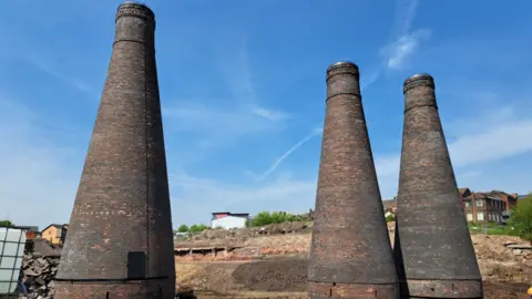 Stoke-on-Trent City Council Three bottle kilns in a line on a building site with houses and trees in the background.