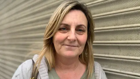 Natalie McNeilly has long blonde hair. She is wearing a grey cardigan and green top. She is standing in front of some shop shutters.