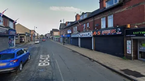 Google Barnsley Road in South Elmsall. A blue car is parked in the front left of the image with a road running away from the camera flanked by rows of shops