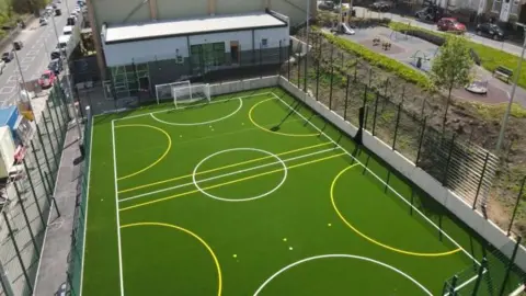 An artists impression of the playzone showing a bright green playing field surrounded by high fences. 