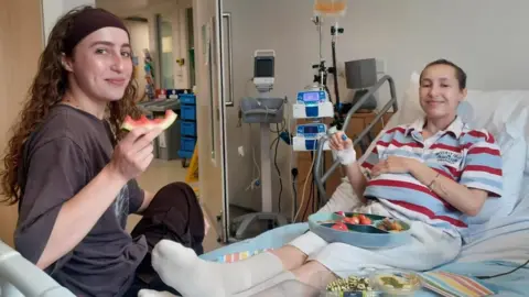 Collect/PA Real Life Grace and Caitlin in hospital. Grace has long dark hair and is wearing a dark top. Caitlin has short hair and is wearing a stripey top