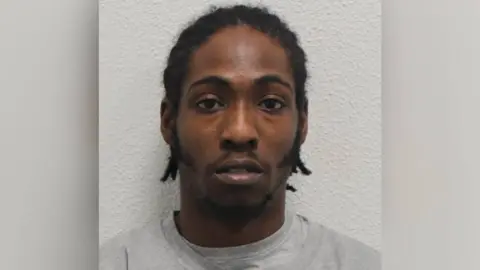 Met Police Custody shot of Omar Peat, wearing grey top and has short black hair in braids, looking straight at the camera.