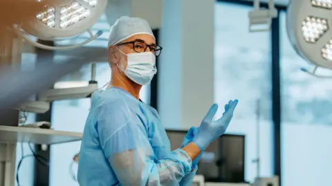 a man in glasses, a hairnet and surgical scrubs pulling a glove onto his hand, in a hospital room setting