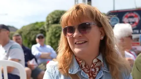 Tracey has long light brown hair and is wearing pink sunglasses. She has a blue jacket and is wearing a white and red shirt. 