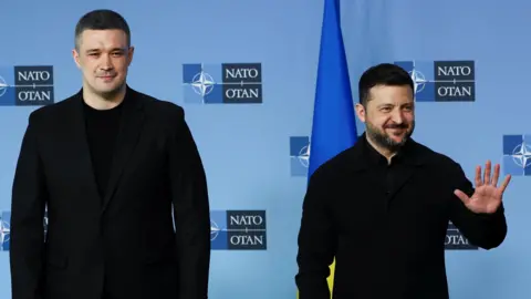 Reuters President Volodymyr Zelensky standing smiling with his hand in a hi-five next to another politician also dressed in black in front of a blue background with the Nato symbol and the Ukrainian flag.
