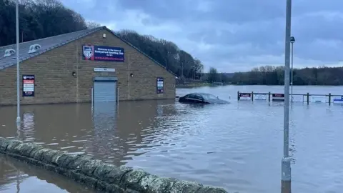 A brick clubhouse building on the left half of the screen, about 15 metres away from the camera. It has flooded to approximately knee-height. A car is to the right of the building and has flooded up to the bonnet. The rest of the image shows flooding. A brick wall near the camera just about pokes above the water. There area trees in the distance.