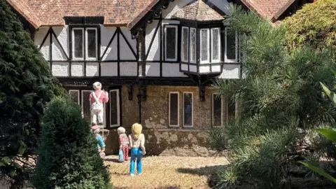 Godshill Model Village A close up of a figure of a topless blonde-haired woman in front of a house with three other model figures working on it.