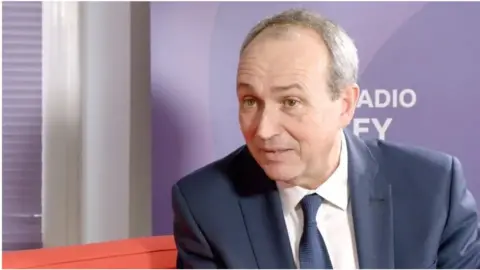 BBC Deputy Lyndon Farnham, Jersey's chief minister, is wearing a blue suit, a blue tie and a white shirt. He is sat on a red sofa in front of a BBC Jersey banner.