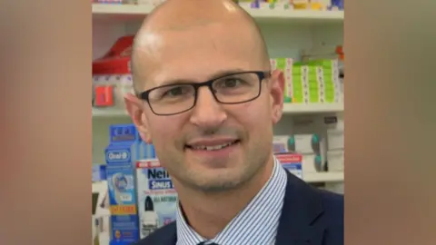 Olivier Picard smiling at the camera, wearing dark framed glasses and a blue stripe shirt with navy blazer.