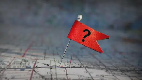 A red flag with a question mark attached to a pin. The pin has been stuck into a map.