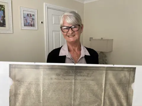 A smiling woman in glasses, a light blouse and dark cardigan holding a large framed historic document